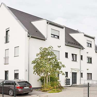 Modernes weißes Mehrfamilienhaus mit dunklem Dach und geparkten Autos davor.