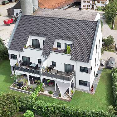Luftaufnahme eines weißen Wohnhauses mit dunklem Dach, Garten und Terrasse neben einer Straße.